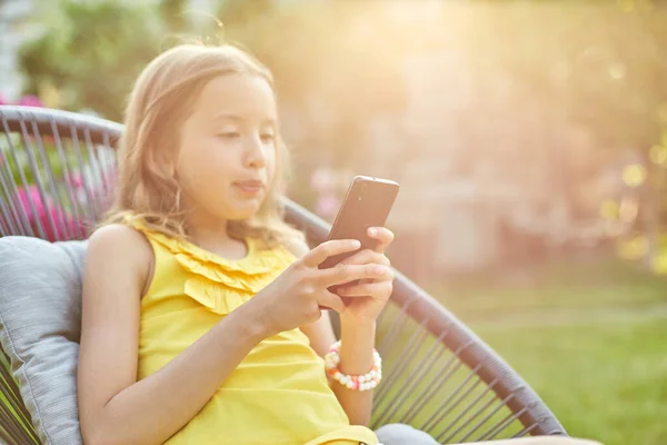 Happy kid girl playing game on mobile phone in the park outdoor, child using smartphone at home garden, backyard, sunlight, smartphone addiction