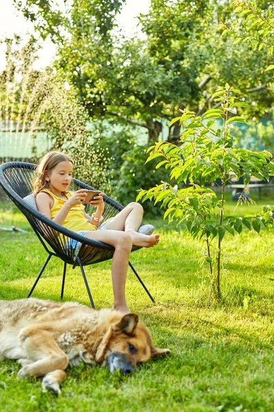 Dog laying on the grass, cute kid girl playing game on mobile phone in the park outdoor, child using smartphone at home garden, backyard, sunlight, Love pets, summer day