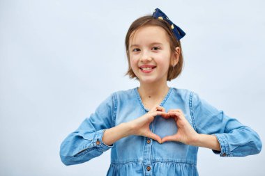 Smiling little girl make heart sign with hands in casual denim dress isolated on white background, studio, makes love icon, gesture with fingers Heart symbol, love concept.