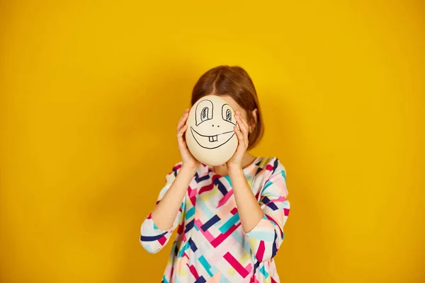 Playful teenager girl covering her face of Ostrich egg with funny face on a bright yellow studio background, Face on the egg, celebrating Easter in style.