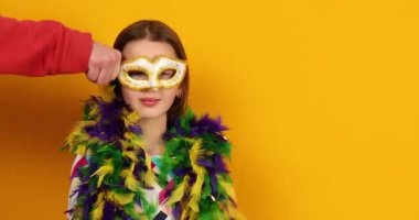 Erkek eli genç kıza renkli bir Brezilya karnaval maskesi takıyor, dans ediyor, sarı stüdyo geçmişinde eğleniyor, kutlamaya hazır, doğum günü partisi, tatil konsepti