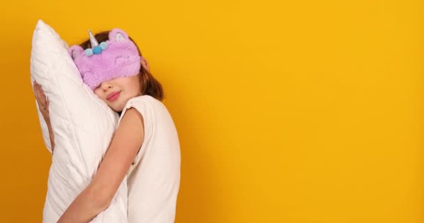 身穿白色睡衣 戴着紫色睡眠面罩的漂亮少女 在工作室里抱着一个黄色背景的枕头 准备睡觉 — 图库视频影像