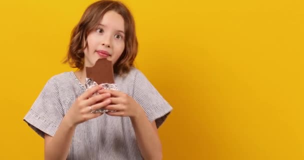 かなり10代の女の子が食べて スタジオの黄色の背景に隔離されたチョコレートバーを噛み デザートを楽しんで スペースをコピー — ストック動画