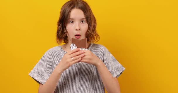 かなり10代の女の子が食べて スタジオの黄色の背景に隔離されたチョコレートバーを噛み デザートを楽しんで スペースをコピー — ストック動画