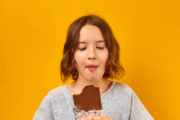 Pretty teen girl eat, bites a chocolate bar isolated on studio yellow background, enjoying dessert, copy space