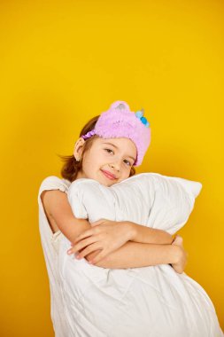 Beyaz pijamalı, menekşe rengi uyku maskeli hoş bir genç kız sarı arka planda, uyumaya hazır bir yastığı kucaklıyor.