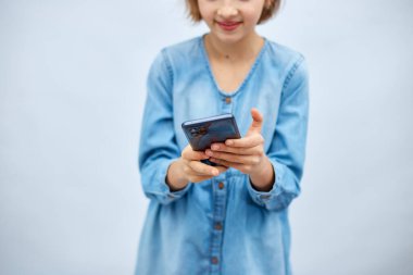 Smiling little girl in casual denim dress hold smartphone, using mobile cell phone internet applications, typing message isolated on white background, studio. Child and modern tech usage concept