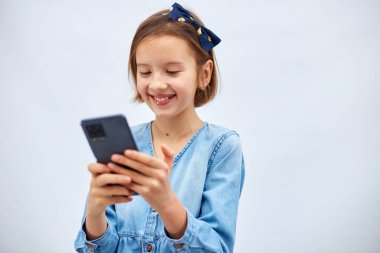 Smiling little girl in casual denim dress hold smartphone, using mobile cell phone internet applications, typing message isolated on white background, studio. Child and modern tech usage concept