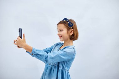 Smiling little girl in casual denim dress hold smartphone do selfie shot on mobile cell phone isolated on white background, studio. lifestyle concept.