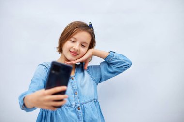 Smiling little girl in casual denim dress hold smartphone do selfie shot on mobile cell phone isolated on white background, studio. lifestyle concept.