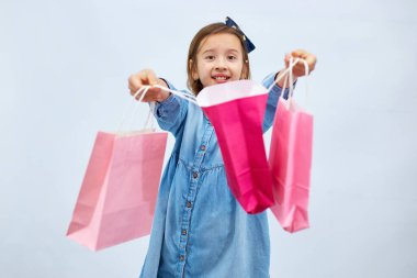 Pretty child girl in casual denim dress hold in hand pink shopping bags on white background, studio, Shopaholic shopping and fashion, discount sale concept, copy space, banner. Black friday
