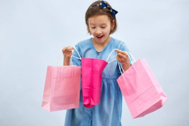 Pretty child girl in casual denim dress hold in hand pink shopping bags on white background, studio, Shopaholic shopping and fashion, discount sale concept, copy space, banner. Black friday, WOW