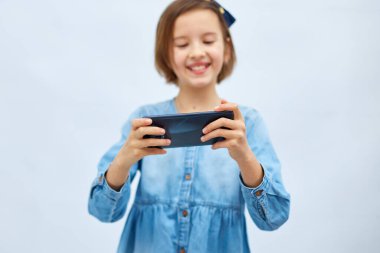 Fun excited kid girl in casual denim dress use with smartphone, enjoys mobile app, internet addiction, play app on mobile cell phone hold gadget isolated on white background, studio