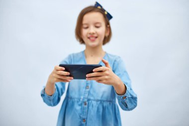 Fun excited kid girl in casual denim dress use with smartphone, enjoys mobile app, internet addiction, play app on mobile cell phone hold gadget isolated on white background, studio