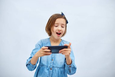 Fun excited kid girl in casual denim dress use with smartphone, enjoys mobile app, internet addiction, play app on mobile cell phone hold gadget isolated on white background, studio