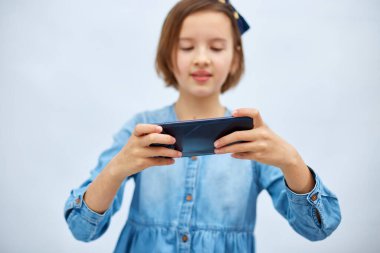 Sad kid girl in casual denim dress use with smartphone, enjoys mobile app, internet addiction, play app on mobile cell phone hold gadget isolated on white background, studio