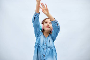 Girl teenager in casual denim dress listening to music in headphones on white background, wireless earphones enjoying music dancing , Modern technology, Protect Developing Ears