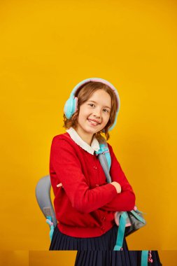 Sırt çantalı mutlu liseli kız, kulaklık takan, tatilde eğlenen okul sarı stüdyo arka planında, okul üniformalı genç kız..