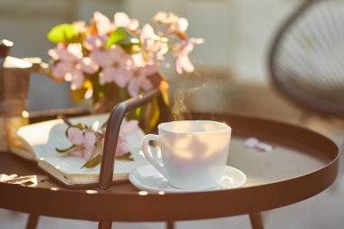 Güneşli bir günde, terasta küçük siyah masanın üzerinde buhar çiçekleri ve defter bulunan bir fincan kahve, açık havada çalışma alanı, yaz tatili, kahve molası..