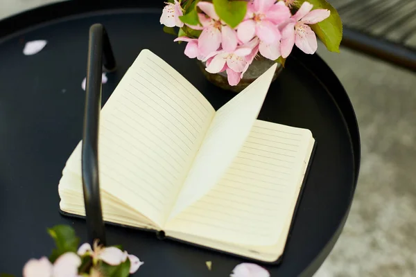 Open notebook and flowers and on the small black table on terrace at home in a sunny day, outdoor workspace, summer relaxation, notes, ideas, plannin