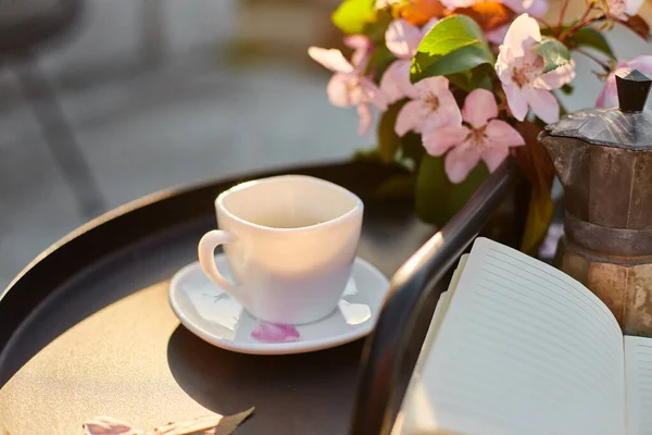 Cup of coffee flowers and notebook on the small black table on terrace at home in a sunny day, outdoor workspace, summer relaxation, coffee break.