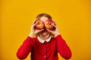 Stüdyonun sarı arka planında, gözlerinin önünde elma tutan zarif, güler yüzlü liseli kız okul konseptine, göz sağlığına geri dönüyor.