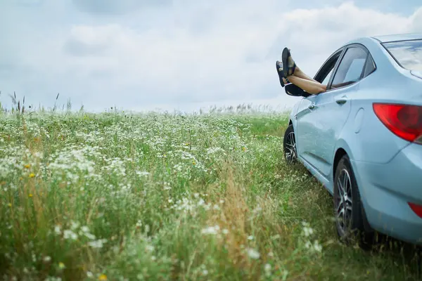 Feet with heels sticking out of a cars window amid a field of white flowers on a cloudy day.