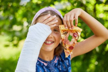 Kolunda beyaz alçı olan neşeli bir kız bir gözünü bir dilim pizzayla kapatıyor..