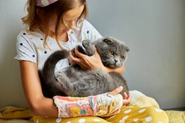 Kırık kollu genç bir kız. Ortopedik alçı. Evde kediyle oynuyor..