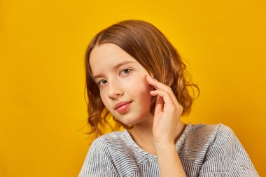 Sakin bir liseli kız, elini yanağında sarı bir zemine koyup düşünceli poz verir..