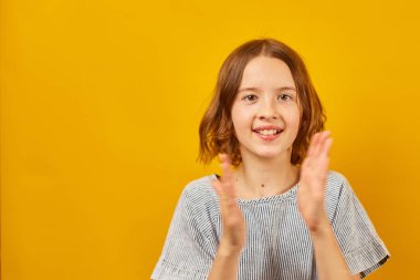 A young girl in a striped shirt claps her hands against a vivid yellow backdrop. clipart