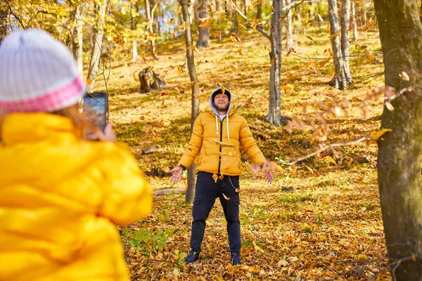 stock image A father joyfully tosses leaves in the air while his daughter captures the moment on her phone, surrounded by vibrant autumn trees.