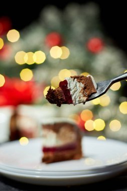 Bir el, güzel katmanlı bir tatlıya doğru nazikçe çatalı kaldırıyor, zenginler çikolata, krema ve canlı meyveler sergiliyor, arka planda tatil süslemeleri var..