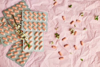 Blister packs with capsules scattered on a textured pink background, alongside small white flowers. clipart