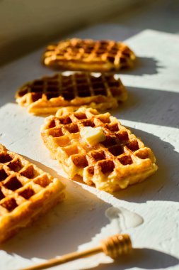 Hafif bir yüzeyde, yanında küçük bir parça tereyağı ve bal kaplayıcısı bulunan waffle 'lar, doğal sabah ışığında lezzetli bir kahvaltı sergiliyor..