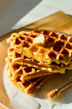 Beyaz bir tabağa istiflenmiş leziz waffle 'lar ve üzerinde küçük bir parça tereyağı eriyor. Yanlara serpiştirilmiş bal ekliyor tatlı bir dokunuş, hepsi de güneş ışığında güzelce aydınlatılıyor..