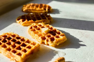 Hafif bir yüzeyde, yanında küçük bir parça tereyağı ve bal kaplayıcısı bulunan waffle 'lar, doğal sabah ışığında lezzetli bir kahvaltı sergiliyor..