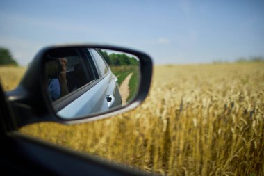 Bir arabanın yan aynası, açık mavi gökyüzünün altındaki altın tarlaların arasında, kırsal alanların güzelliğini gösteren dolambaçlı toprak bir yol görüntüsü yakalar..