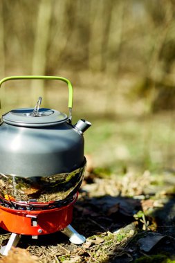 Çaydanlığı olan taşınabilir bir soba ormanda yerde oturur, güneş ışığı ağaçların arasından süzülür. Çaydanlık ısıtma suyudur, açık hava yemekleri veya çay için idealdir..