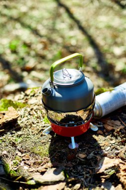 Çaydanlığı olan taşınabilir bir soba ormanda yerde oturur, güneş ışığı ağaçların arasından süzülür. Çaydanlık ısıtma suyudur, açık hava yemekleri veya çay için idealdir..