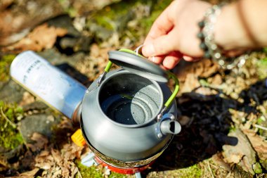 Bir insan, sakin bir ormanla çevrili taşınabilir bir ocakta su kaynatmaya hazırlanıyor. Güneş ışığı ağaçların arasından süzülür, açık hava deneyimini arttırır..