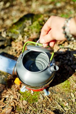 Bir insan, sakin bir ormanla çevrili taşınabilir bir ocakta su kaynatmaya hazırlanıyor. Güneş ışığı ağaçların arasından süzülür, açık hava deneyimini arttırır..