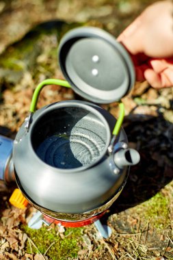 Bir insan, sakin bir ormanla çevrili taşınabilir bir ocakta su kaynatmaya hazırlanıyor. Güneş ışığı ağaçların arasından süzülür, açık hava deneyimini arttırır..