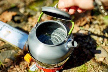 Bir insan, sakin bir ormanla çevrili taşınabilir bir ocakta su kaynatmaya hazırlanıyor. Güneş ışığı ağaçların arasından süzülür, açık hava deneyimini arttırır..