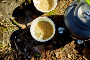 Bir grup arkadaş dışarıda rahatlar, kağıt bardaklardan sıcak içecekler yudumlarlar. Parlak güneş ışığı, yosunlu zemindeki bir su ısıtıcısının yanında buluşmalarını aydınlatır..