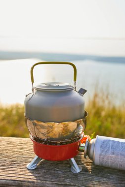 Güneş arka planda batarken sakin bir gölün yakınındaki portatif bir ocağa bir çaydanlık yerleştiriliyor. Ilık parıltı, sakin atmosferi güçlendirir. Açık hava yemekleri için idealdir..