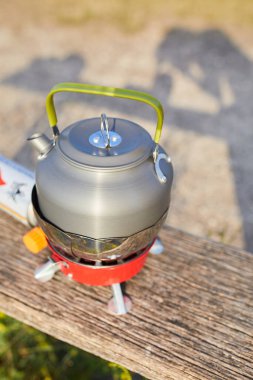 Güneş arka planda batarken sakin bir gölün yakınındaki portatif bir ocağa bir çaydanlık yerleştiriliyor. Ilık parıltı, sakin atmosferi güçlendirir. Açık hava yemekleri için idealdir..