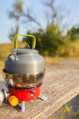 Güneş arka planda batarken sakin bir gölün yakınındaki portatif bir ocağa bir çaydanlık yerleştiriliyor. Ilık parıltı, sakin atmosferi güçlendirir. Açık hava yemekleri için idealdir..