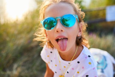 Parlak güneş ışığı, parlak güneş gözlüğü takarken dilini şakacı bir şekilde uzatan genç bir kızı aydınlatıyor. Yeşil arka plan, dışarıda geçirdiği neşeli ana neşe katıyor..