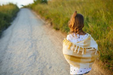 Genç bir kız çamurlu bir yolda yürüyor, yaz güneşinin tadını çıkarıyor. Yeşil tepeler ve doğa ile çevrili, omuzlarının üzerine açık bir şal ve kot şort giyiyor..